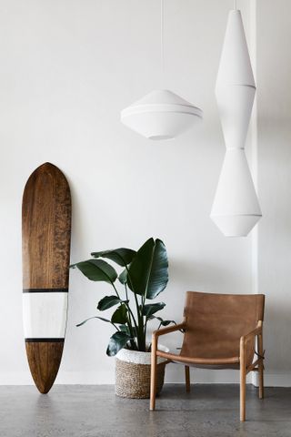 Interior view of a space at Studio Warrior One featuring white walls, grey floors, white pendant lights, a brown chair, a green plant in a woven pot and a wooden, black and white surfboard leaning against the wall