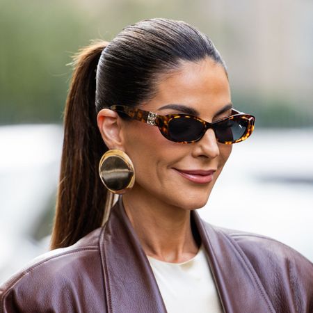 A woman wearing a burgundy leather jacket, white tee, sunglasses and has a slicked back ponytail hairstyle