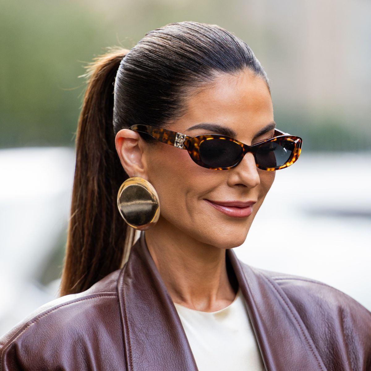A woman wearing a burgundy leather jacket, white tee, sunglasses and has a slicked back ponytail hairstyle
