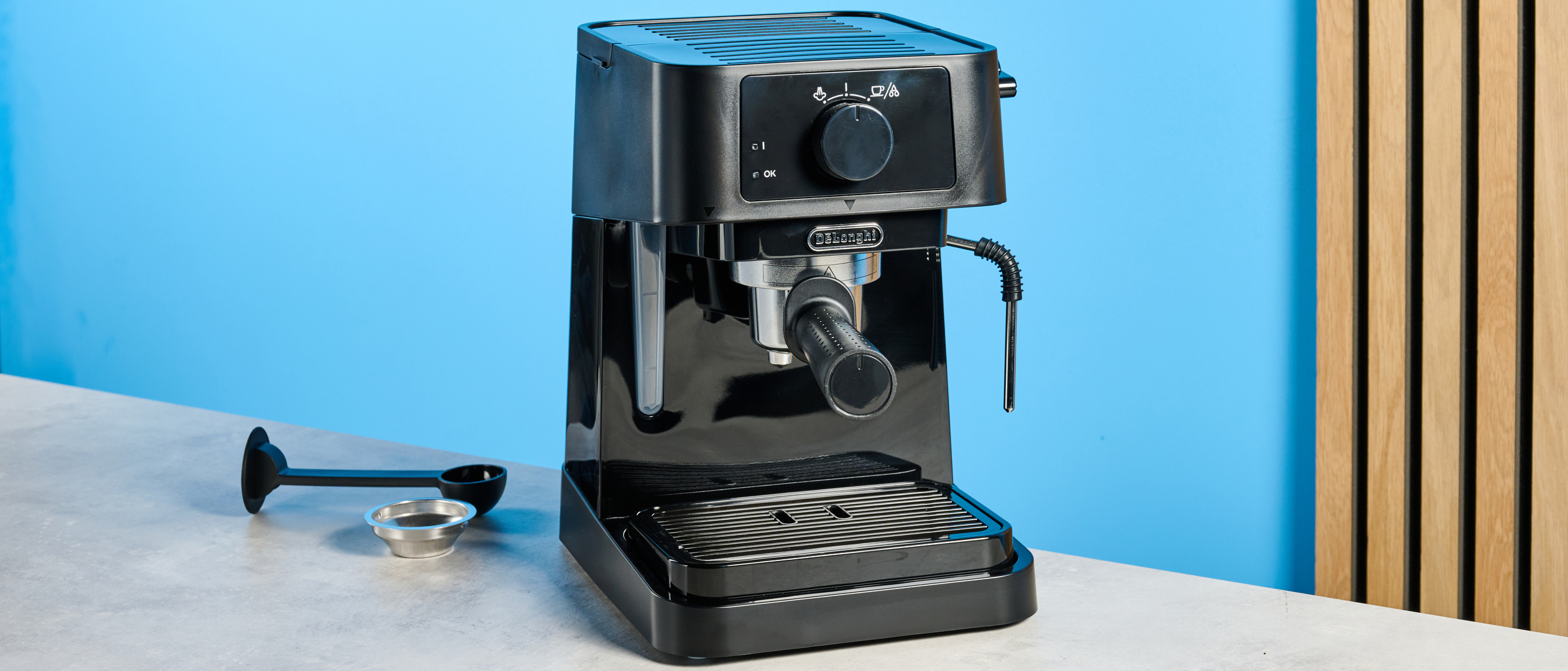 the de'longhi stilosa espresso machine in black, with one portafilter, a dial for espresso and milk steaming, and a plastic tamper, photographed against a blue background