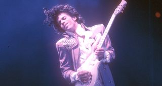 Prince plays a solo on his white Cloud guitar onstage during his 1985 Purple Rain Tour, bathed in white and purple light.