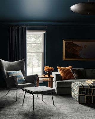 Image of a dark living room that is painted in a teal blue color. There is a gray accent chair with an ottoman in front of it.