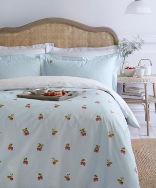 A light blue bed with a red strawberry pattern, with a light brown jute headboard, a wooden tray with tomatoes and bread on the bed, and white scalloped nightstand with a glass vase, white tea pot, and books underneath