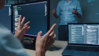 A close-up shot from the back of a cybersecurity worker speaking with a colleague who is standing across from them, with code visible on a laptop and set of monitors on the original worker&#039;s desk. Decorative: Neither person&#039;s head is in the frame and both are wearing business attire. The shot is dimly-lit.