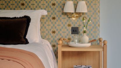Bedroom with statement geometric wall, light wood nightstand, gold sconce lamp with white lampshades, and bed with white bedding, brown cushion, and peach blanket.