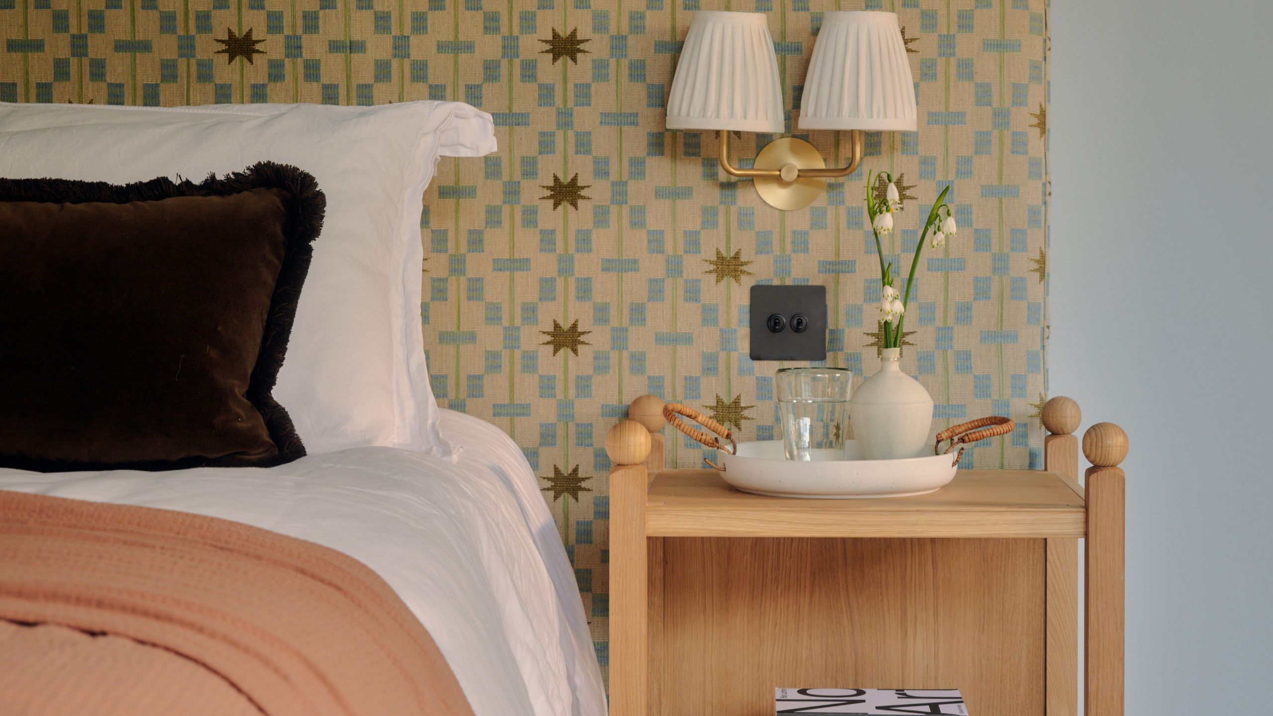 Bedroom with statement geometric wall, light wood nightstand, gold sconce lamp with white lampshades, and bed with white bedding, brown cushion, and peach blanket.