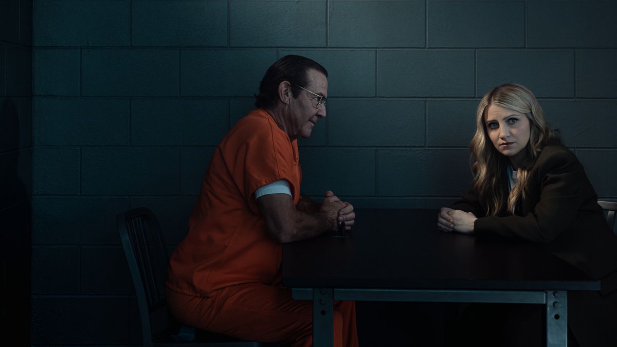 Convicted killer Keith Hunter Jesperson (Dennis Quaid), wearing an orange prison jumpsuit, sits opposite his wearied daughter Melissa Moore (Annaleigh Ashford) in a visitation room in a still from the true crime drama series Happy Face.