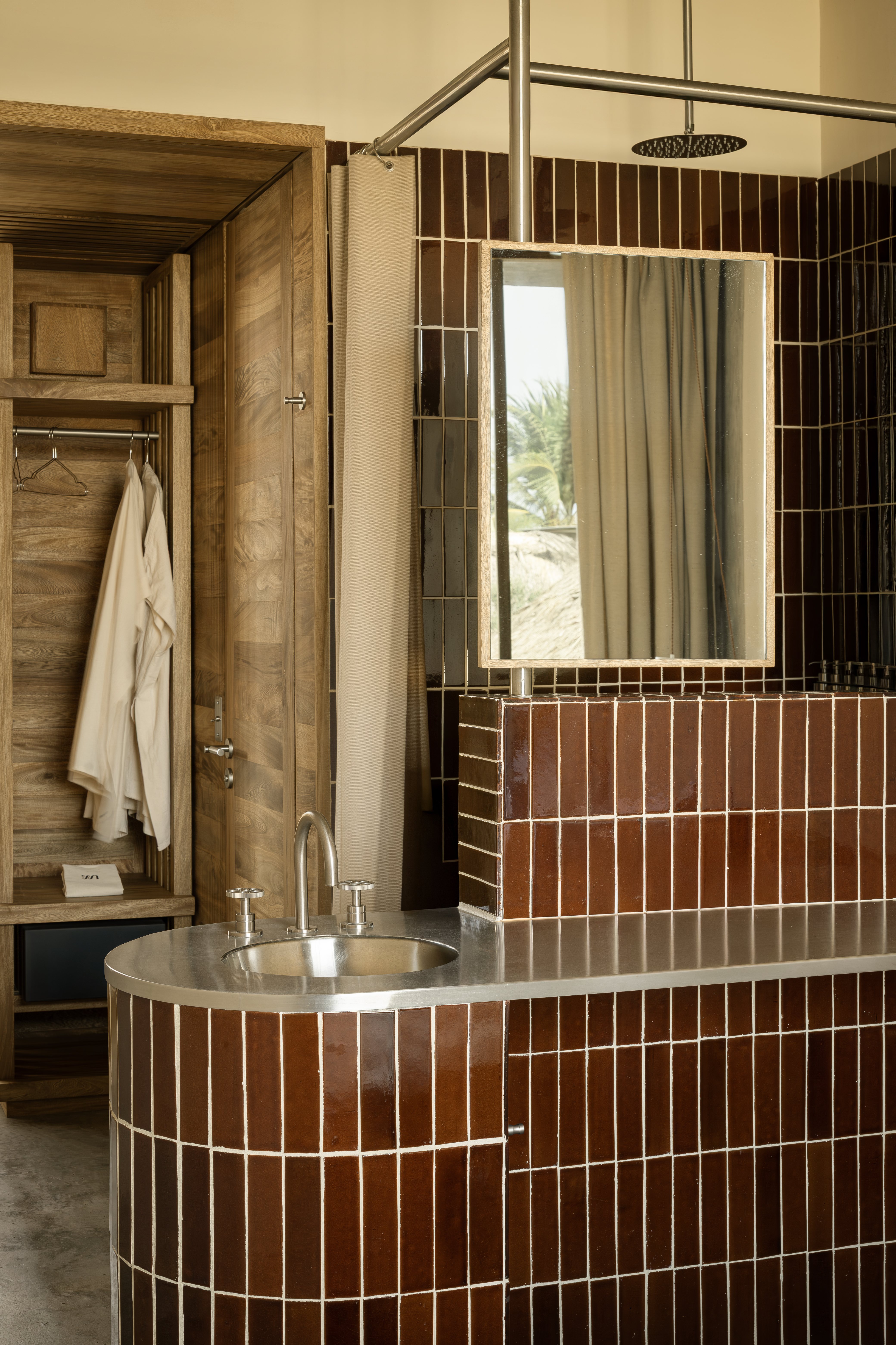 A brutalist hotel sculpted from wood and cement features brown tile-clad bathrooms with chrome sink tops, linen towels, and wooden-framed mirrors.