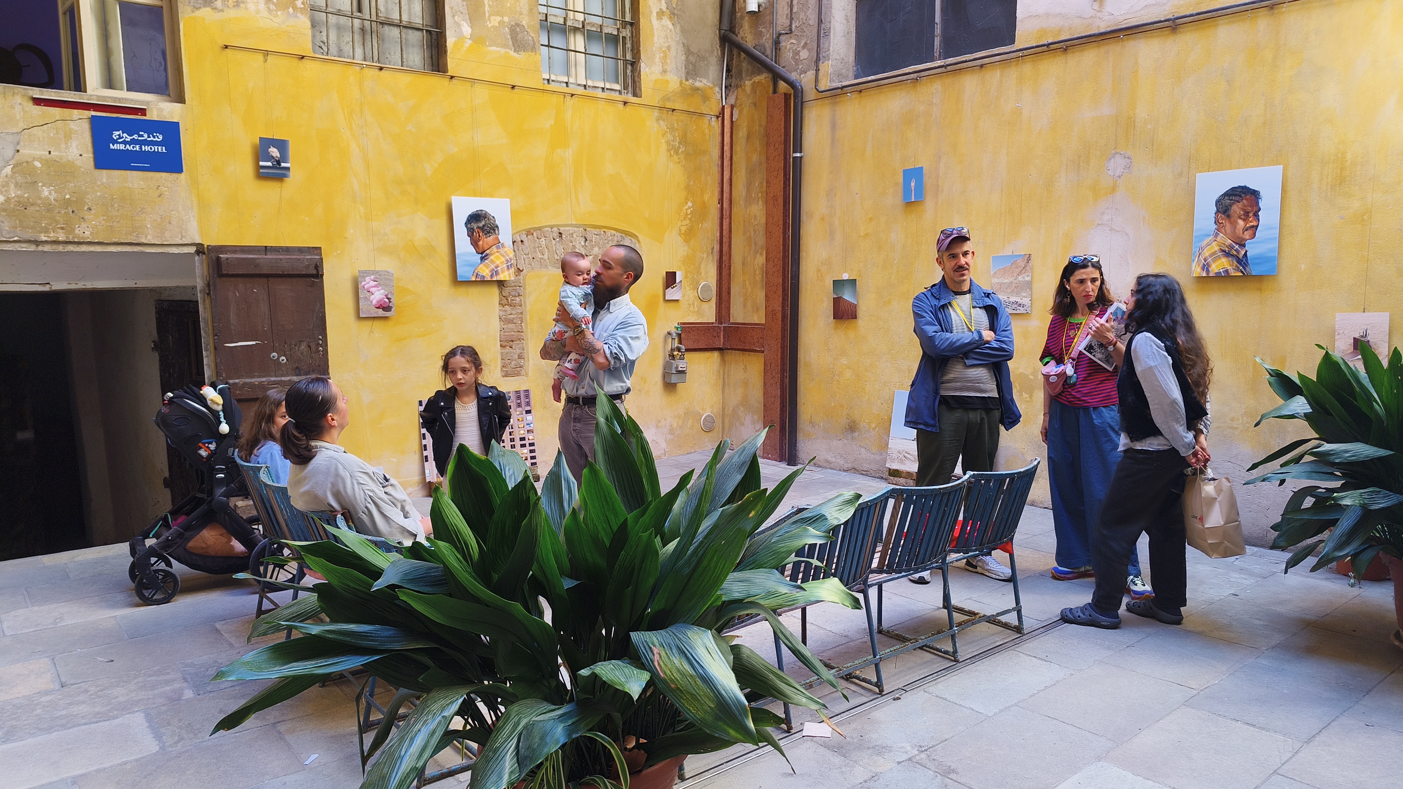 Visitors gather in a sunlit yellow-walled courtyard at the Mirage Hotel exhibition, with small colour photographs mounted on the walls and a tattooed man cradling a baby in the foreground.