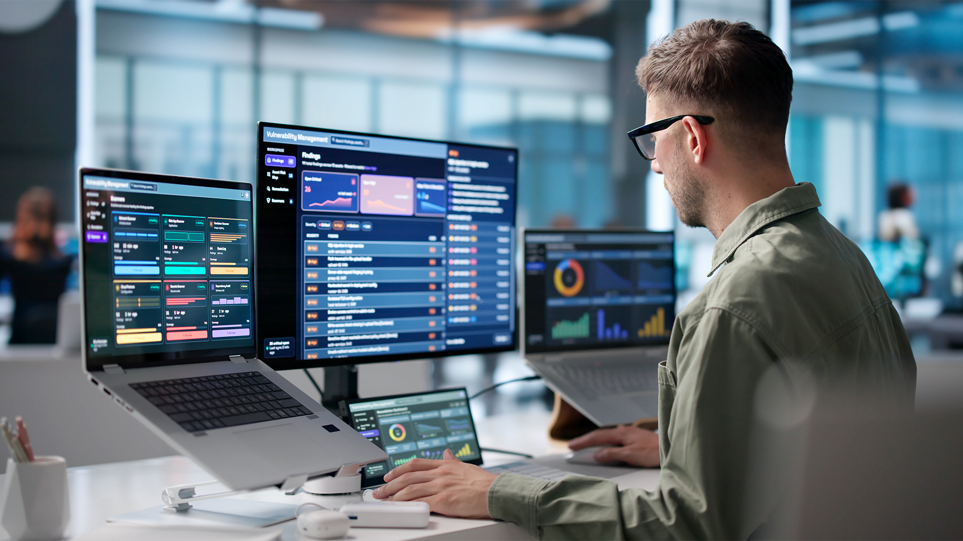Male cybersecurity expert conducting penetration test on company network while working on a desktop computer with laptops attached to monitor, with vulnerability management portal on screen.