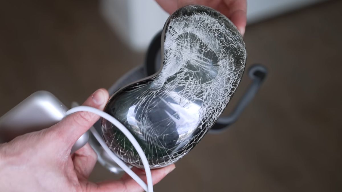 A person picks up a broken Vision Pro, which has a cracked front glass panel.