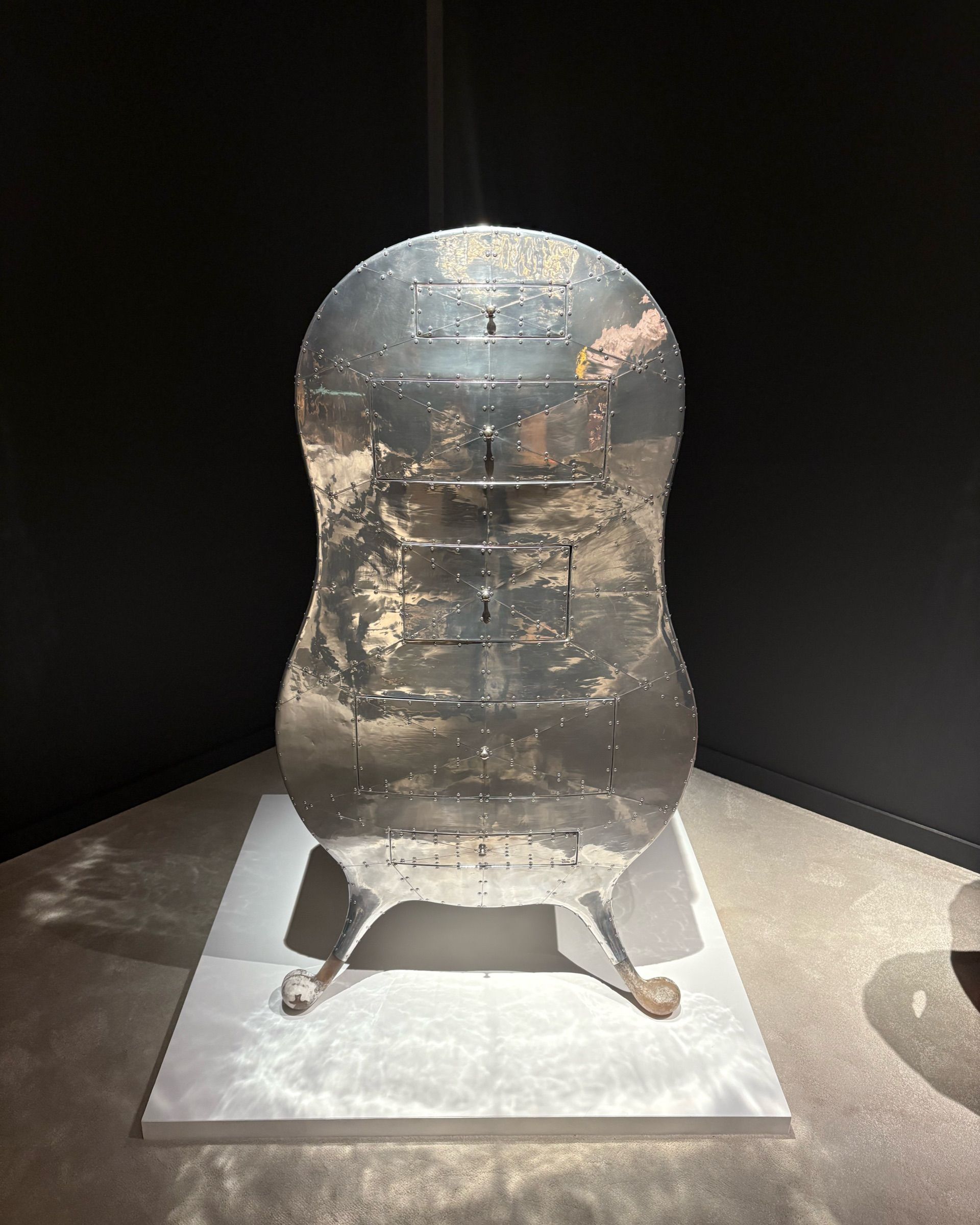 A silver cabinet with a violin-shaped body, a textural, reflective surface, and creaturesque legs stands on a white podium positioned atop a beige carpet.