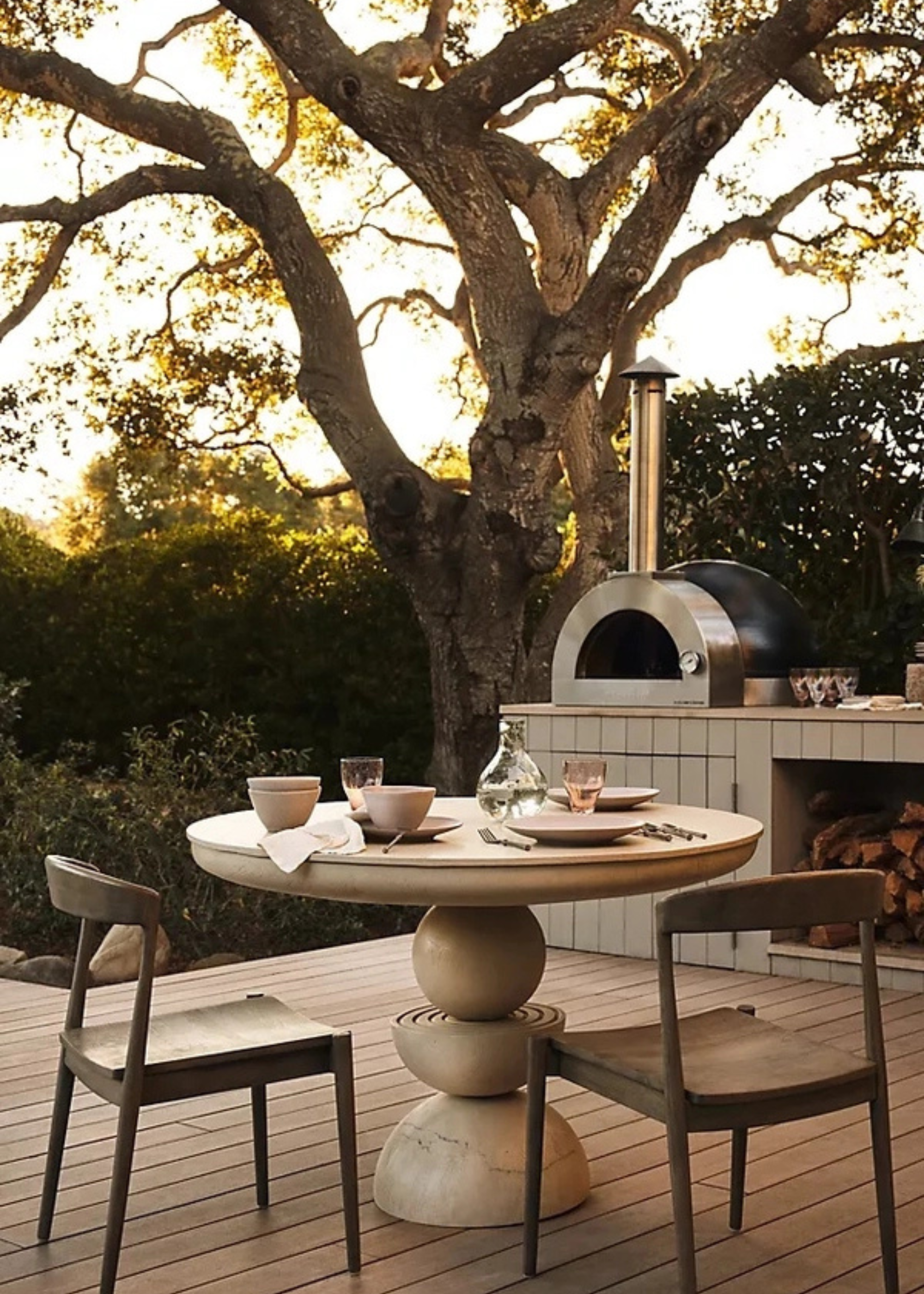 a modern outdoor dining table with a built-in pizza oven in the background next to a large tree