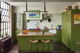 A green beadboard kitchen cabinets with a kitchen island and bar stools. There is a bright orange stove and oven place as well as hanging pots and pans in the space.