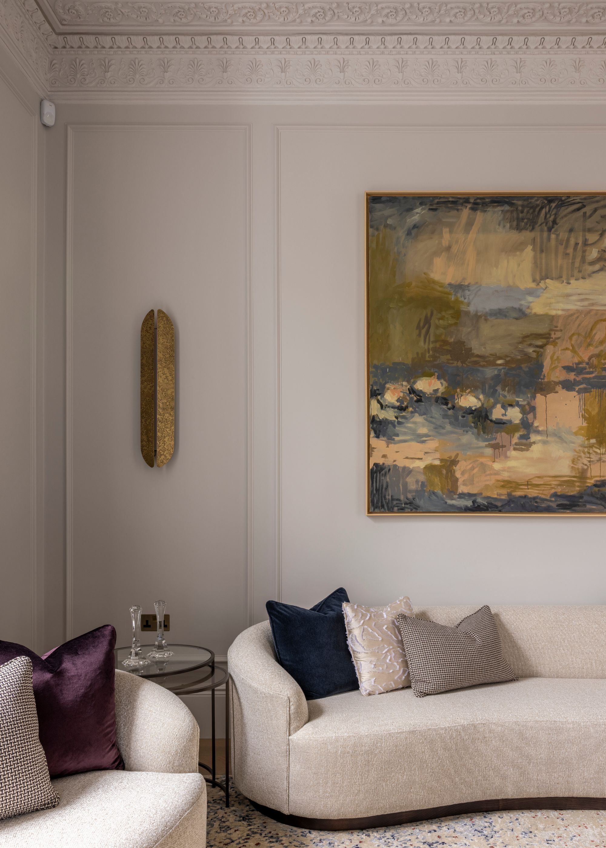 Living room in a traditional property with tall ceilings and ornate coving. Pale gray walls with an abstract artwork in muddy colors, a low curved sofa, and jewel-toned cushions.