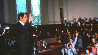 Jim Jones makes a peace sign for his congregation from the pulpit in California