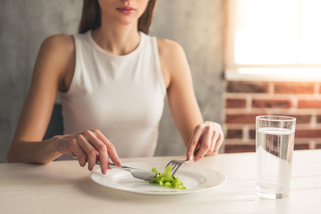 Cropped image of a woman eating a single piece of broccoli with a glass of water next to her plate. 