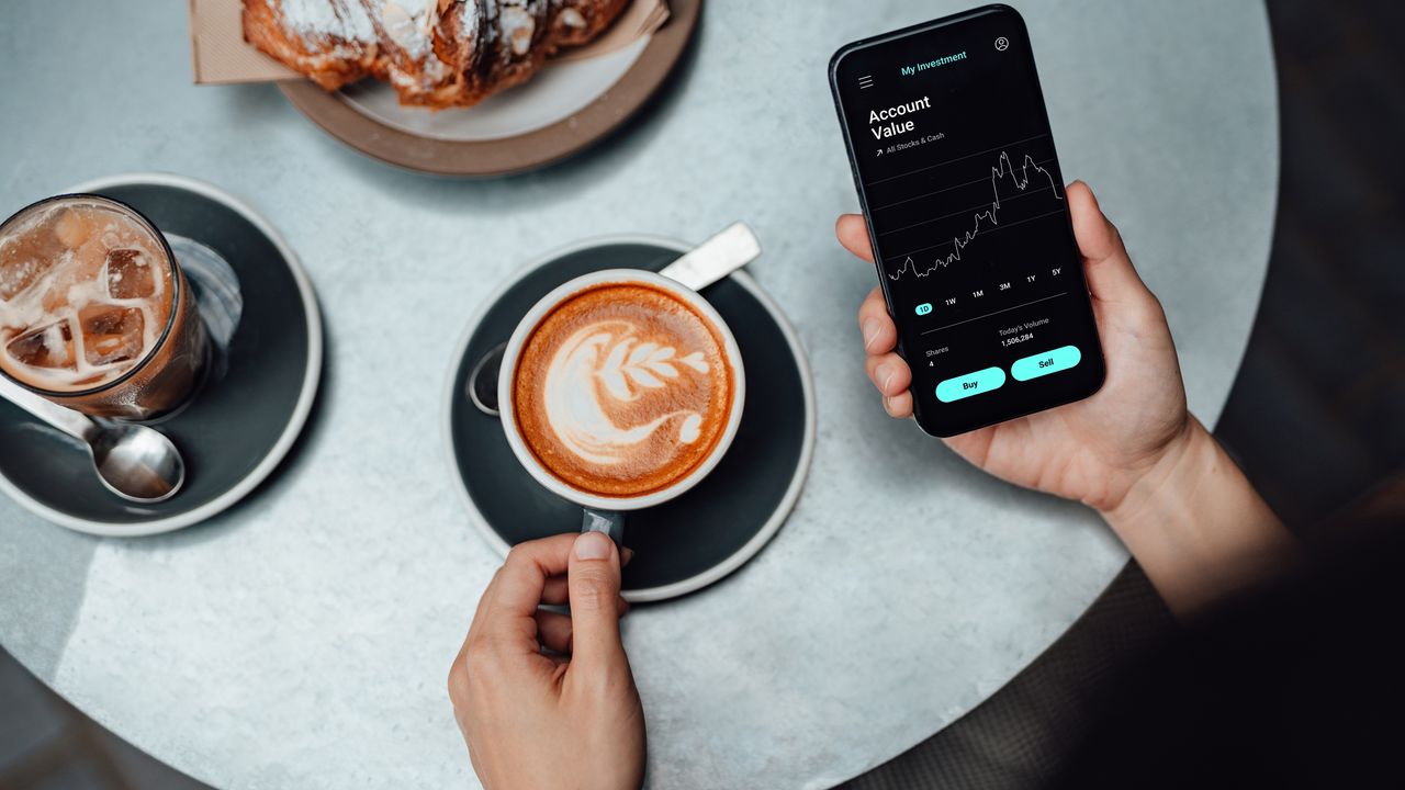 An unseen person picks up a cup of cappuccino while looking at market trading on a smartphone.