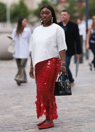 Nnenna Echem wearing a white asymmetric round-necked t-shirt with a knee-length red skirt embroidered with red sequins, and red mesh ballet flats