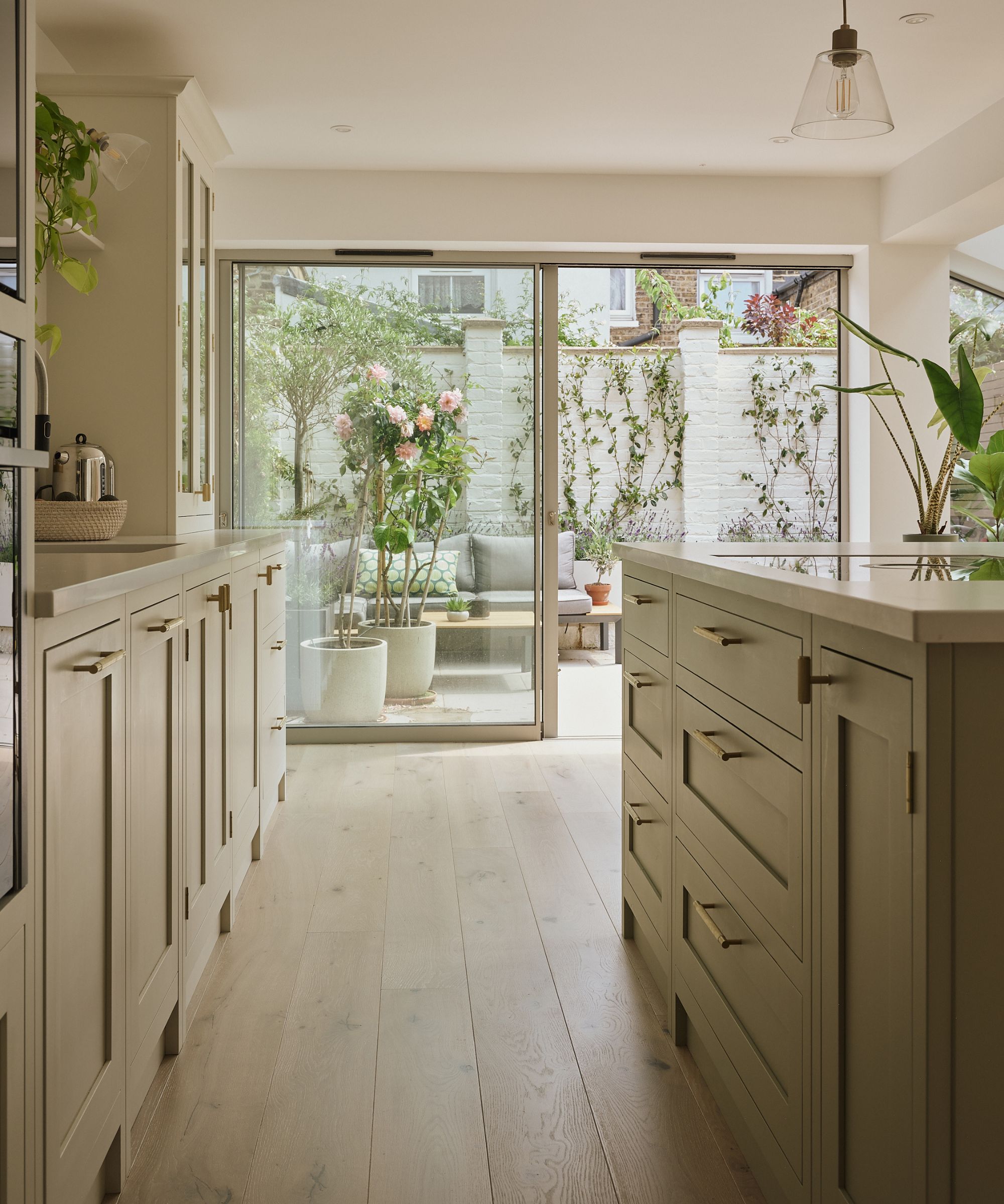 A very light sage green kitchen with gold cabinet handles. A real light pine wooden floor. Large sliding glass doors in the background with a view onto a patio with lots of potted trees.