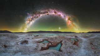 Panoramic view of the Milky Way arcing over a salt flat with turquoise pools and distant mountains at twilight
