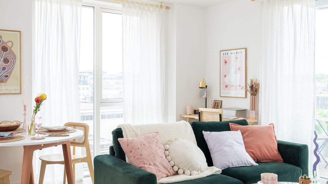 A small living room with a dark green velvet sofa, a dining area and floor-length voile curtains on big windows