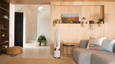 Clean, brown living room with Dyson tower fan in the centre, part of a gray sofa with bright cushions to the right, a shelf with plants and a scenic painting, a bookshelf to the left, a foot stool, and an opening leading to stairs and a door.