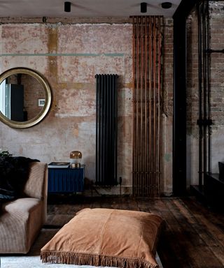 Living room with exposed brick and plaster wall, wooden floor, copper pipes floor to ceiling and tall black radiator, large floor cushion on rug beside armchair.