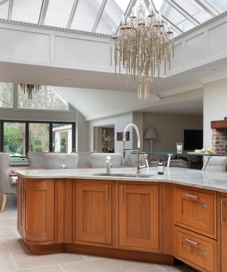 A white kitchen area with a white glass ceiling with a chandelier hanging from it and a kitchen island with gray granite surfaces, a basin and faucet, and warm wooden cabinets