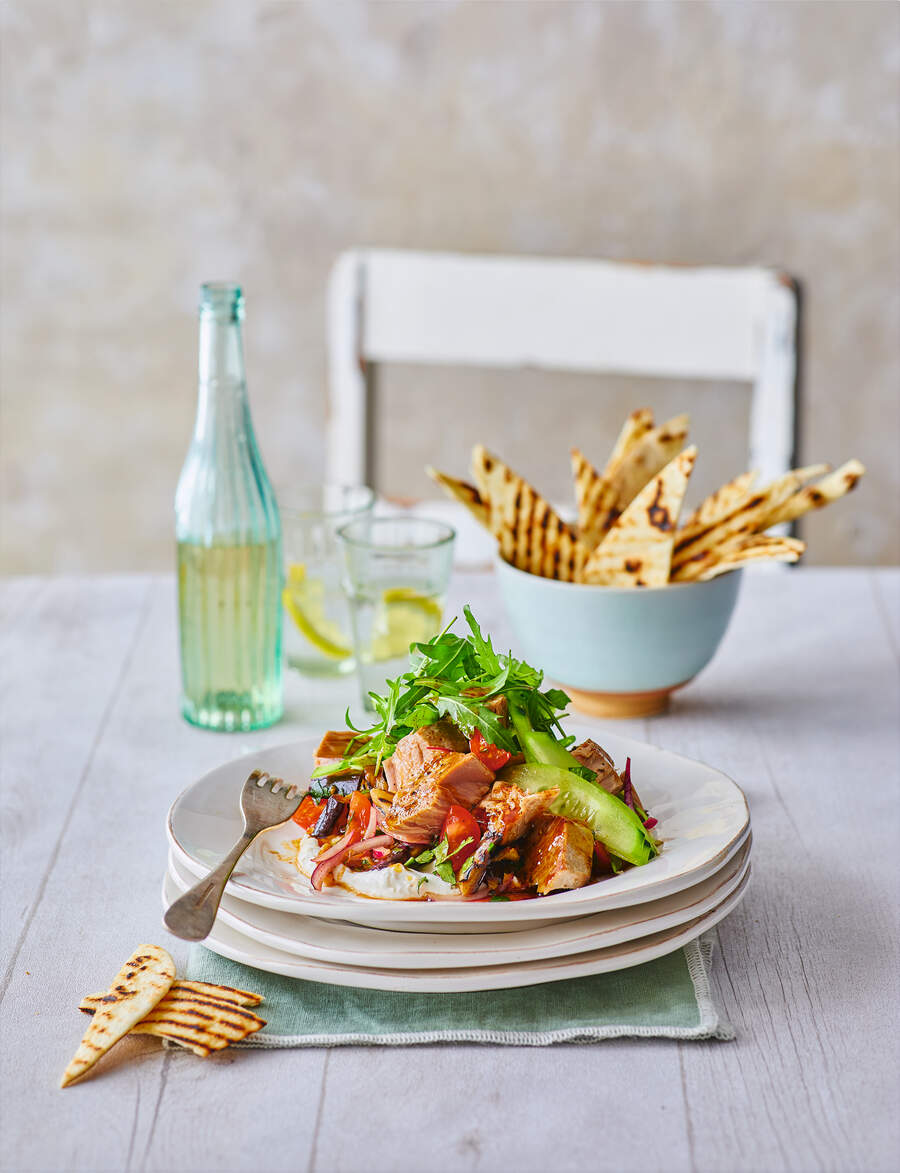 A plate of freshly cooked tuna flakes on a tangy aubergine salad, garnished with fresh vegetables, accompanied by a glass of lemon-infused water and a bowl of grilled flatbreads.