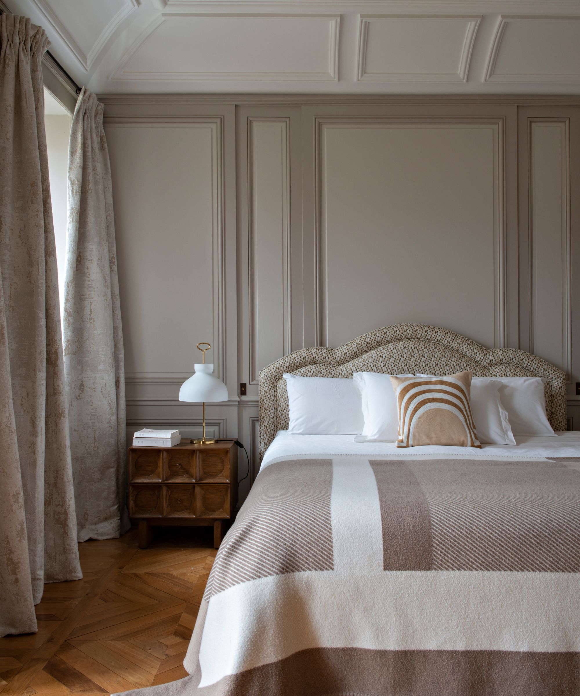 Bedroom with walls painted in Farrow &amp;amp; Ball's Elephant's Breath, parquet wooden flooring, an upholstered headboard, white sheets and a beige quilt, and a wooden nightstand with a glass lamp on top.