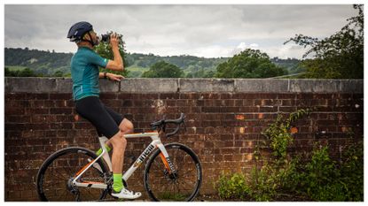 A male cyclist leans against a wall while drinking from a water bottle