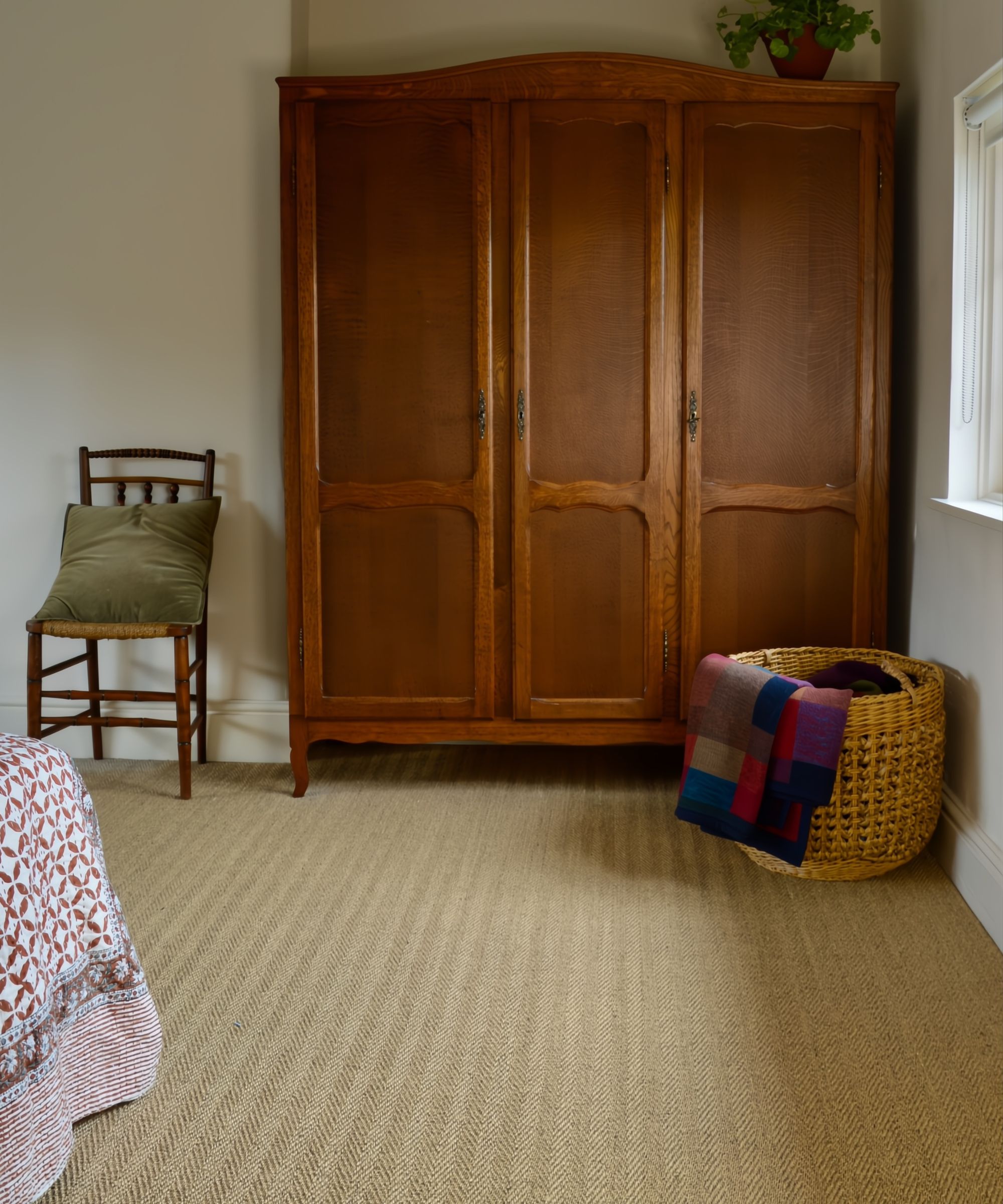 A large antique armoire wardrobe with three doors on a sisal striped carpet. A wash basket sits in front with a colourful throw spilling out. A small chair sits to the left of the wardrobe with an olive green velvet cushion