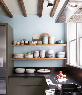 dark grey green kitchen cabinets with pale blue walls and open wooden shelves with beamed ceiling above