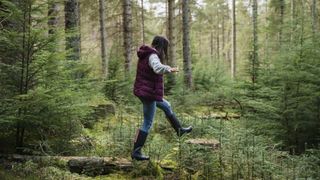 A woman in outdoor gear walks through a pine forest. She is in the process of stepping from a felled log to a tree stump with her arms outstretched.