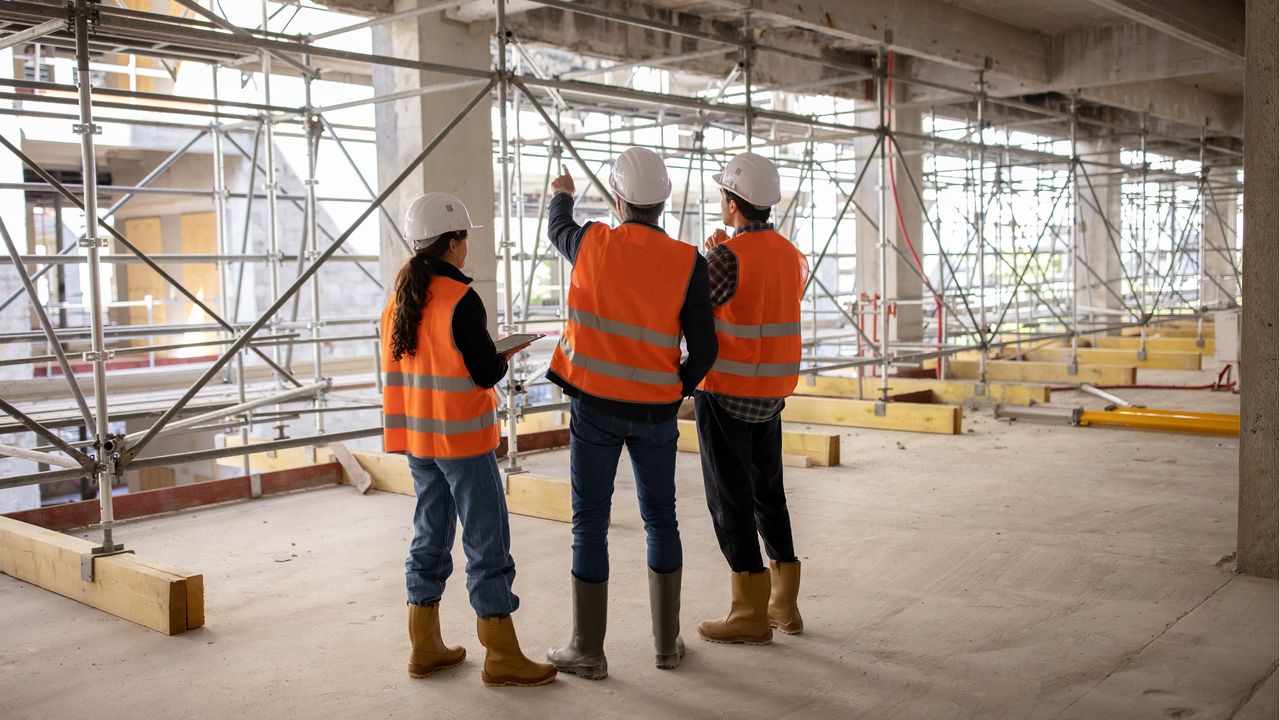 A woman and two men wearing orange safety vests stand in a building that&#039;s under construction.