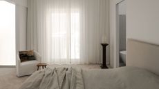 A white bedroom with sheer white curtains, a white sofa chair with a patterned pillow, a wood stool, a pedestal with a vase, and a bed with a beige headboard, and gray bedding beside a bathroom with a tub