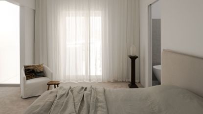 A white bedroom with sheer white curtains, a white sofa chair with a patterned pillow, a wood stool, a pedestal with a vase, and a bed with a beige headboard, and gray bedding beside a bathroom with a tub
