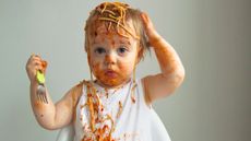 A toddler holding a fork has spaghetti all over her head, face and bib.