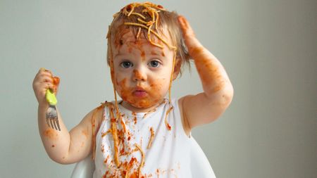 A toddler holding a fork has spaghetti all over her head, face and bib.