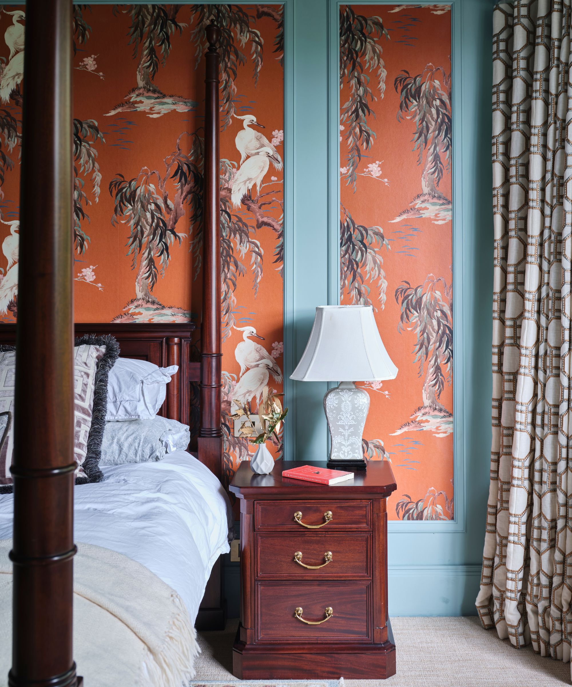 A bedroom with blue walls with panelled orange statement wallpaper, a dark wood four poster bed with white bedding with a matching bedside table with white lamp, patterned curtains, and neutral carpet.
