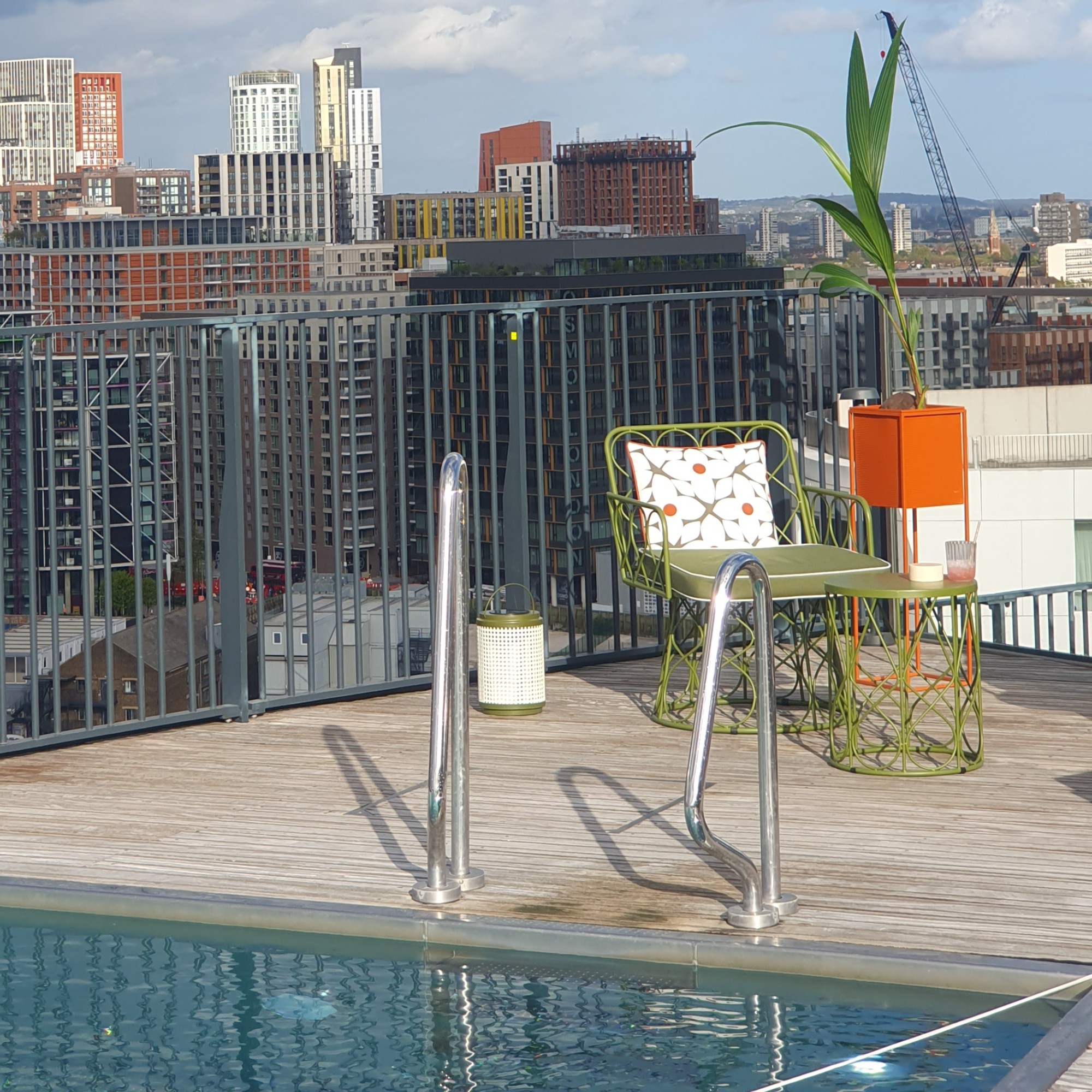 A rooftop pool with QVC's Studio 70 metal garden chair in green metal with a matching side table and a square orange planter next to it
