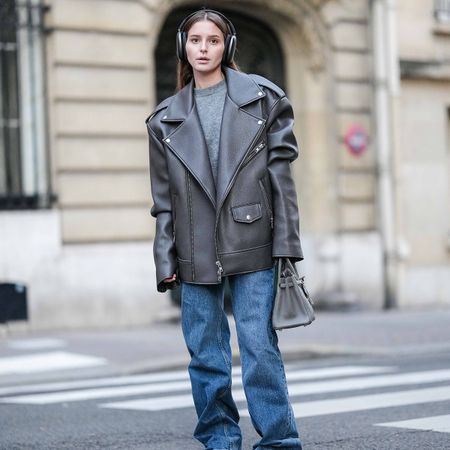 A woman wears a gray leather jacket, gray sweater, puddle jeans, a gray hermes bag, and gray ugg boots.