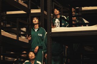 A group of people wearing matching green tracksuits stand between metal bunk beds, in 'Squid Game' season 2.