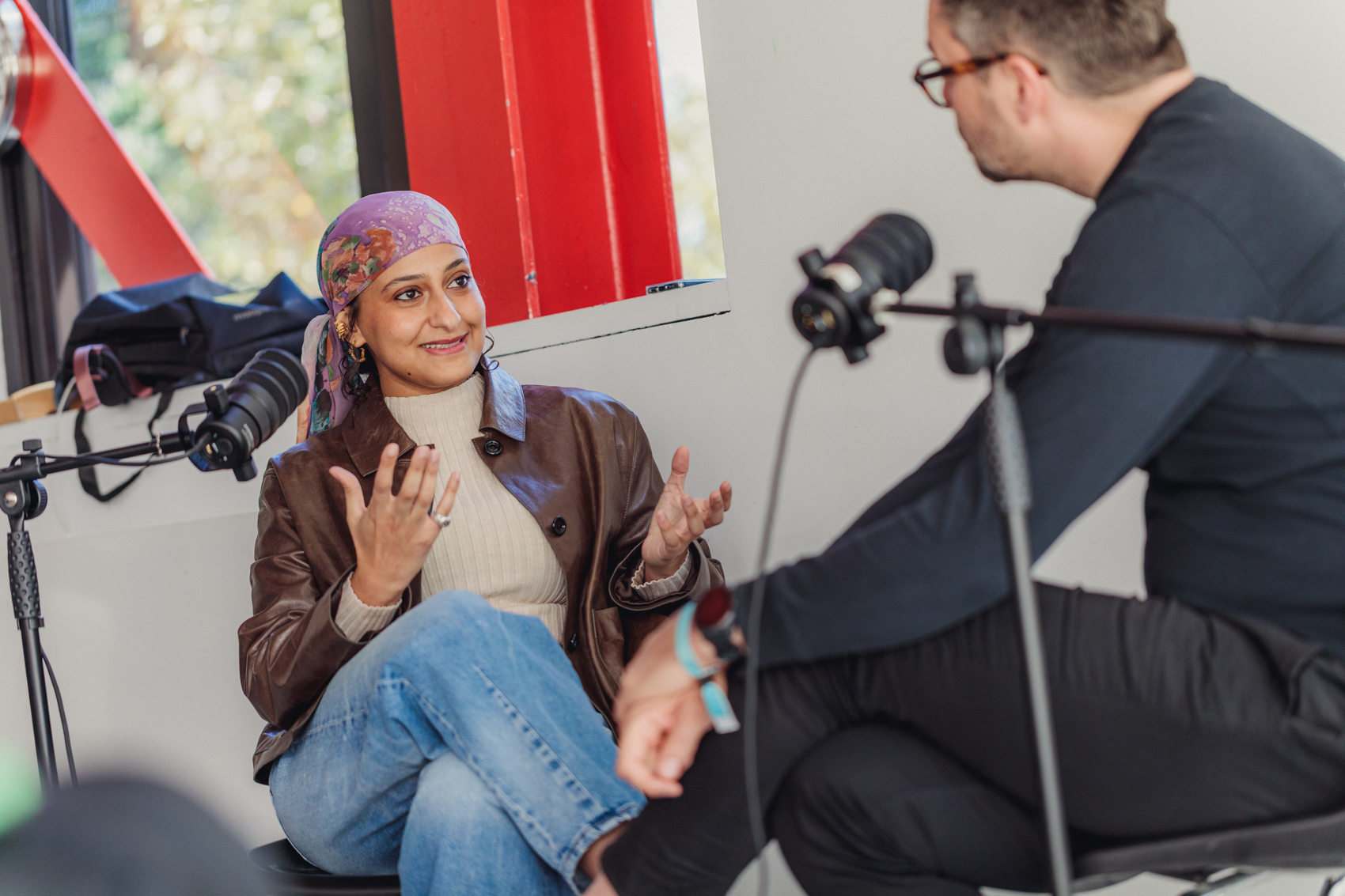 A woman wearing a purple headscarf and brown leather jacket gestures while speaking into a microphone during a podcast or interview with a man wearing glasses.