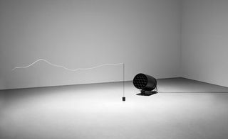 A string of merino wool fluttering from a metal pole being blown by a black metal floor fan an in an all white room