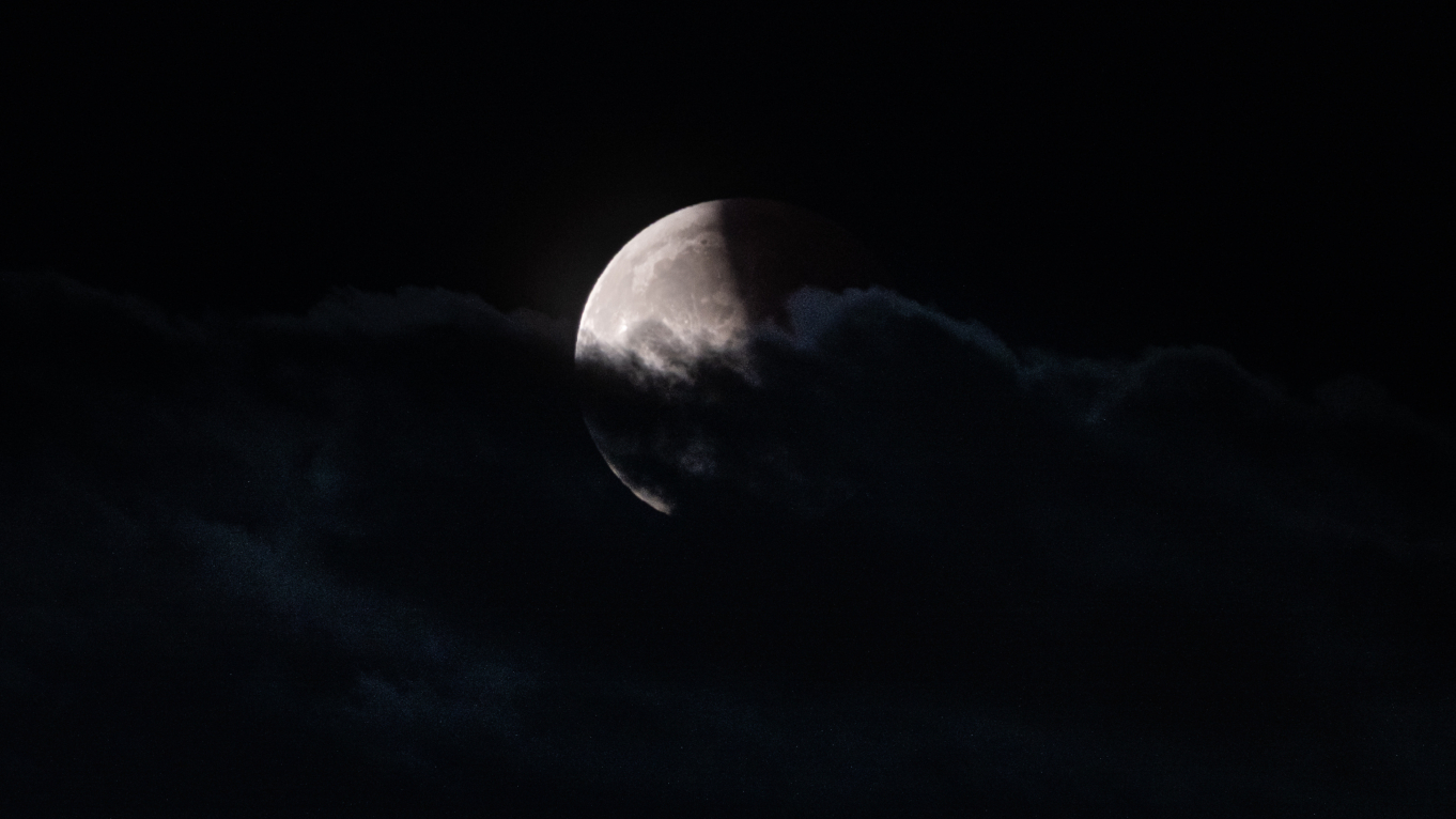 A full moon is pictured during a lunar eclipse with its right side bathed in Earth's shadow against a black sky. Clouds cover the lower section of the lunar disk.