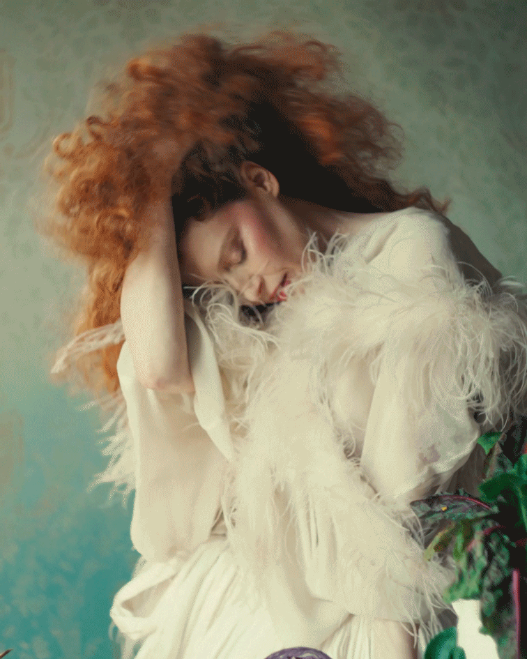 moving gif of Natasha Lyonne pushing back her red curly hair wearing a feathered cape