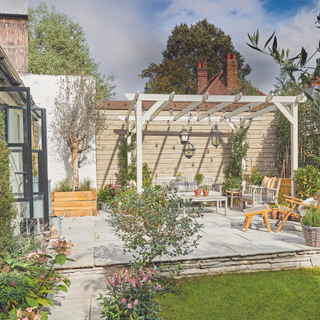 Patio area with wooden table, chairs and bench and plants under white wooden pergola in garden