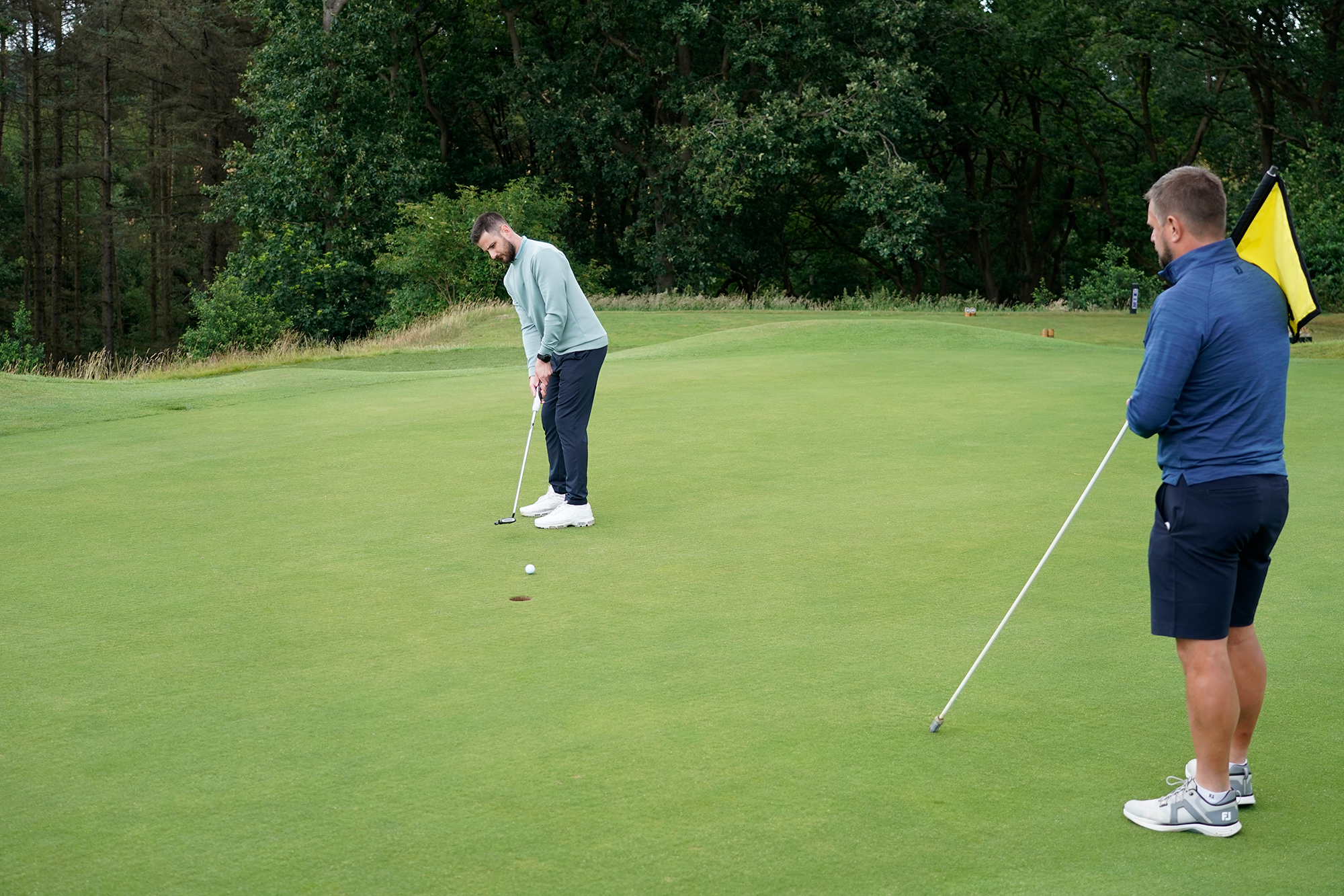 Baz Plummer hitting a putt on the green with Alex James coaching and holding the flag near the hole